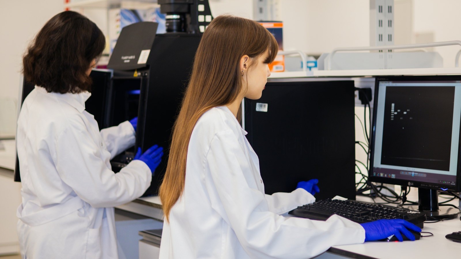 Two researchers in lab coats and gloves working at instruments in a laboratory.